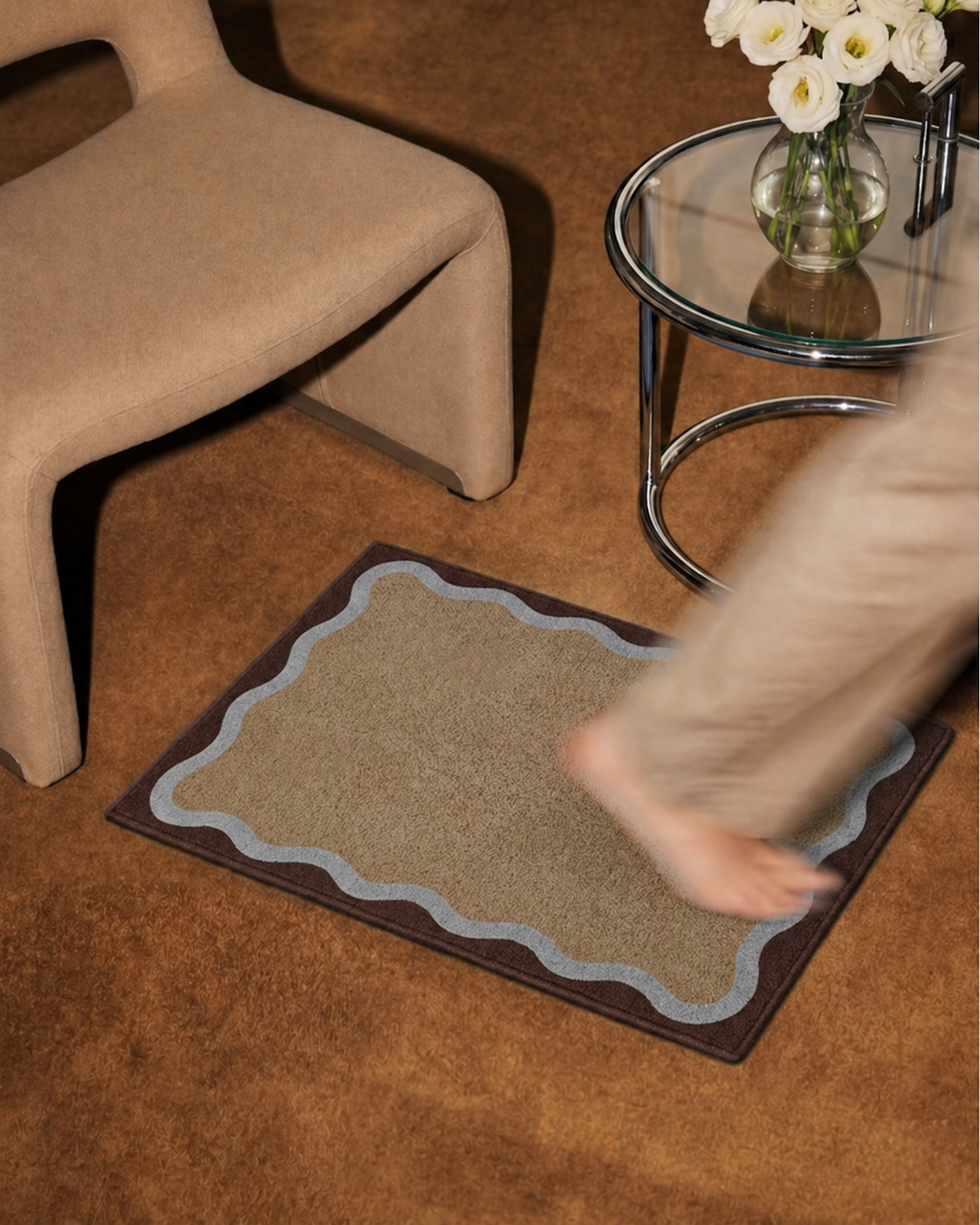 HADDEN Beige rug with scalloped edge on a brown floor next to a chair and glass table with flowers.