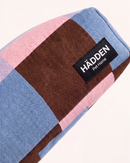 A close-up of a checkered fabric pet bed or blanket by HÄDDEN Pet Home, featuring blocks of light blue, pink, and brown on a white background.
