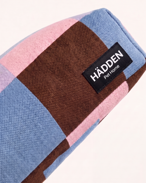 A close-up of a checkered fabric pet bed or blanket by HÄDDEN Pet Home, featuring blocks of light blue, pink, and brown on a white background.
