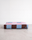 A rectangular dog bed with a checkered pattern in pink, light blue, and dark brown, resting on a light gray concrete floor against a white wall. A small black brand tag for 'HADDEN' is visible on the bottom right.