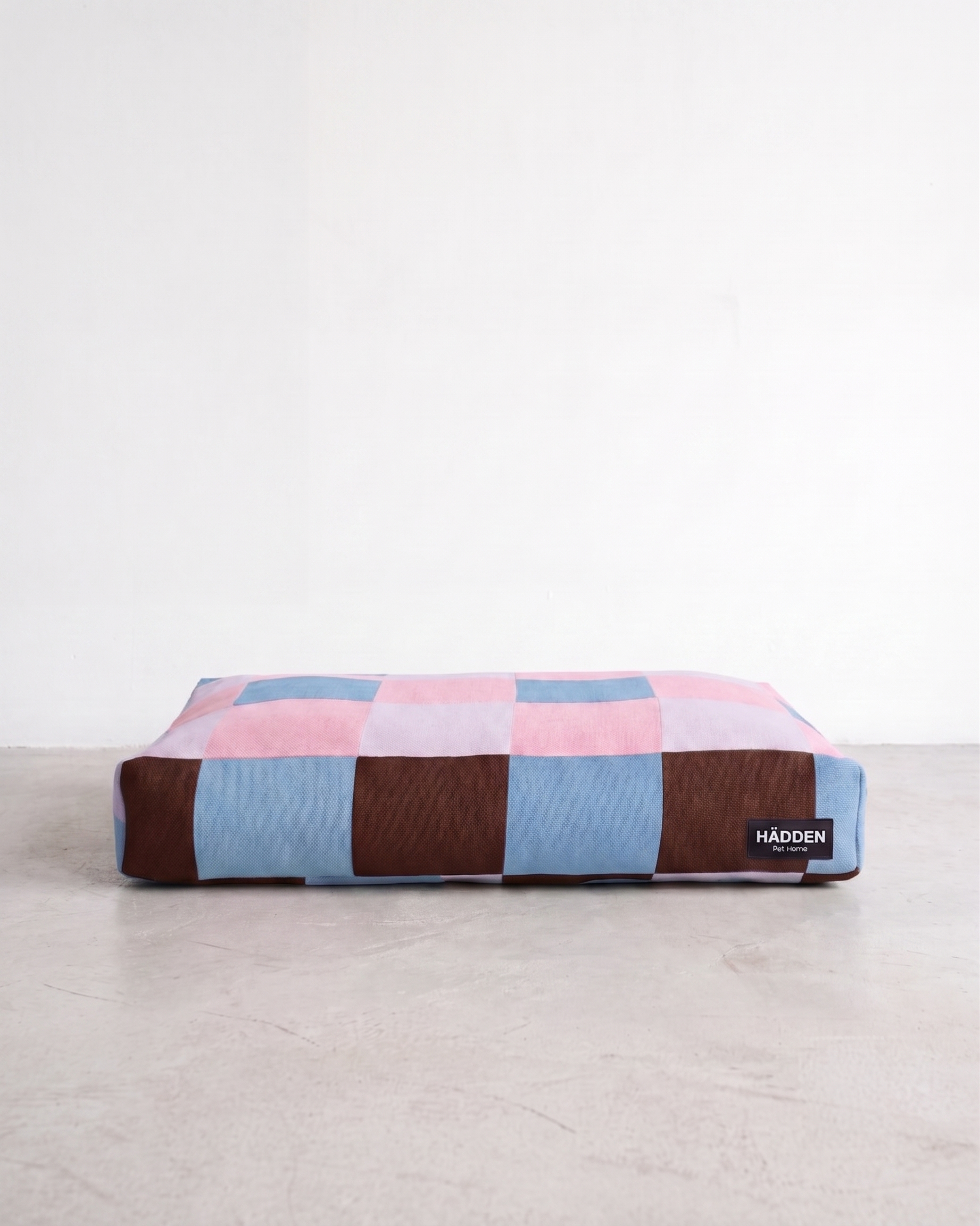 A rectangular dog bed with a checkered pattern in pink, light blue, and dark brown, resting on a light gray concrete floor against a white wall. A small black brand tag for 'HADDEN' is visible on the bottom right.